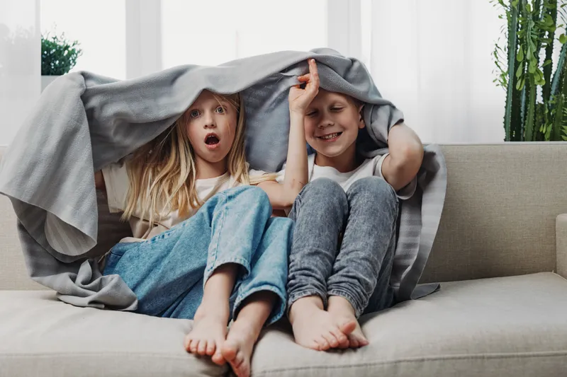 Two happy children sitting on a sofa covered with a blanket, enjoying playful moments at home. Cozy atmosphere with natural light and casual clothing in a living room setting.