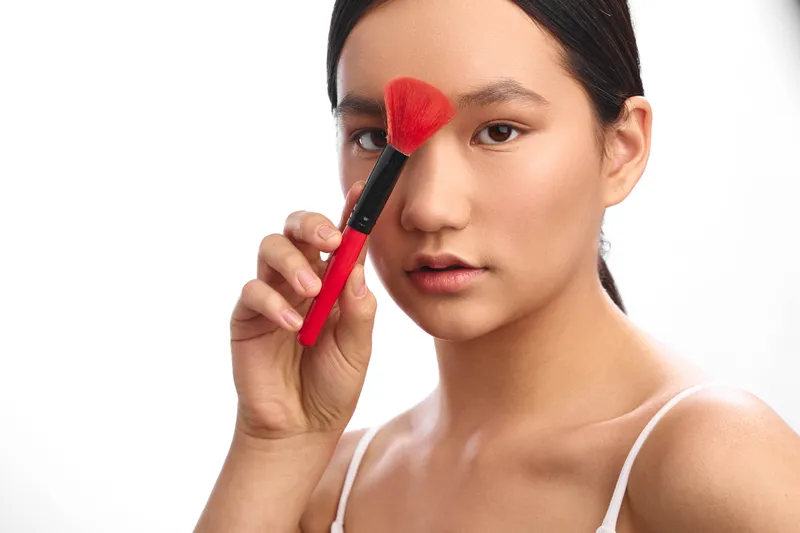 Asian woman applies makeup with a soft brush in a bright studio, showcasing calm focus and graceful beauty as she prepares her skin for a flawless look.