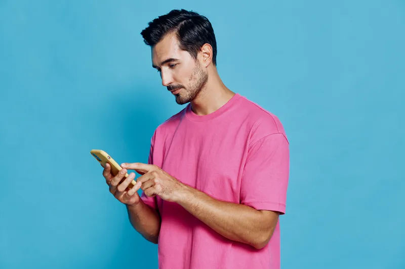Young smiling man wearing a pink t-shirt focused on his smartphone while standing against a solid blue background, expressing a modern lifestyle concept.
