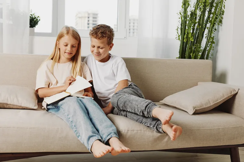 Two children sitting on a beige sofa reading a book together in a bright living room with natural light and plants. Concept of learning, childhood, friendship, and family time.