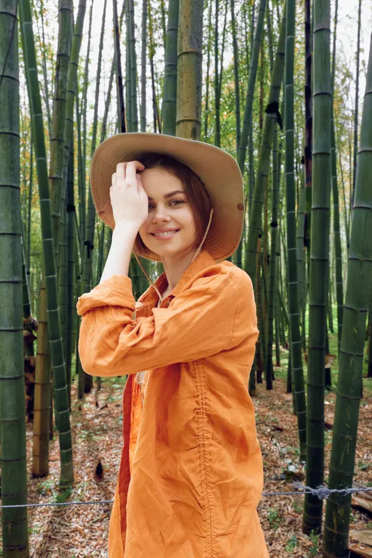 Woman in orange jacket and wide brim hat with a warm smile in a dense bamboo forest, portrait of a traveler enjoying nature, casual pose, vertical composition and serene outdoor mood.