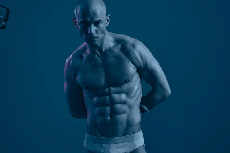 Muscular man posing confidently in a studio setting, showcasing strength and determination. The image has a blue light tone, creating a dramatic atmosphere.