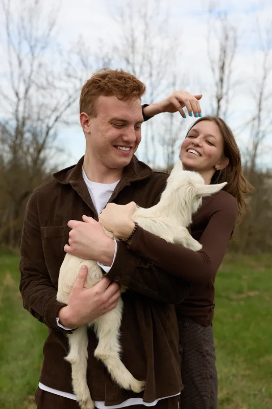 A joyful couple outdoors cradle a playful goat kid, sharing a lighthearted moment in a grassy field. A candid lifestyle scene emphasizing warmth, companionship, and the bond between people and