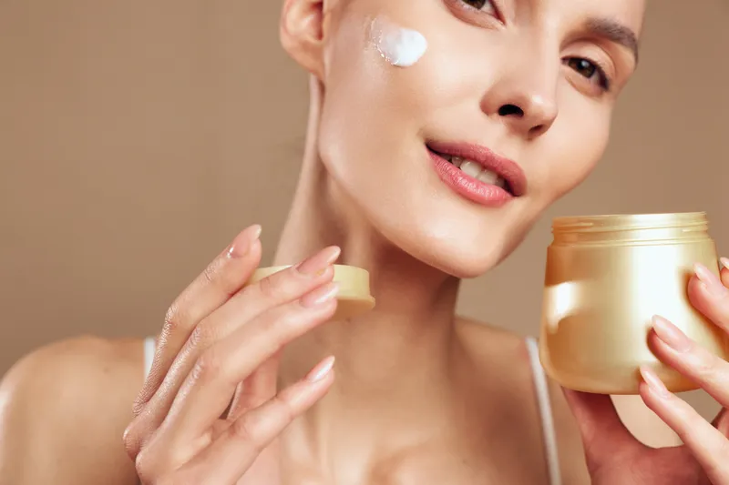 A young woman applies moisturizing cream on her face with a finger, holding an open jar of skincare product. The close-up captures smooth skin and a neutral background, concept for beauty care.
