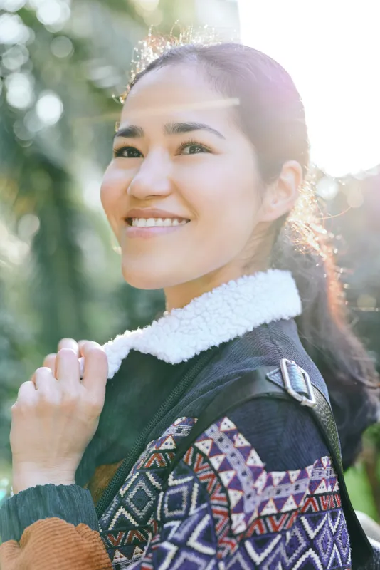 Smiling woman outdoors wearing a patterned knit sweater, warm sunlight and natural greenery setting, showcasing relaxed style, cheerful mood, and casual fashion.