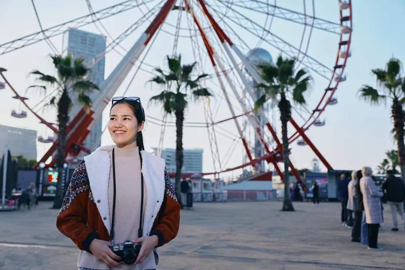 Woman holding camera stands before a large ferris wheel at an outdoor plaza, smiling, casual jacket, daytime cityscape, travel photography vibe, candid street scene, cheerful moment