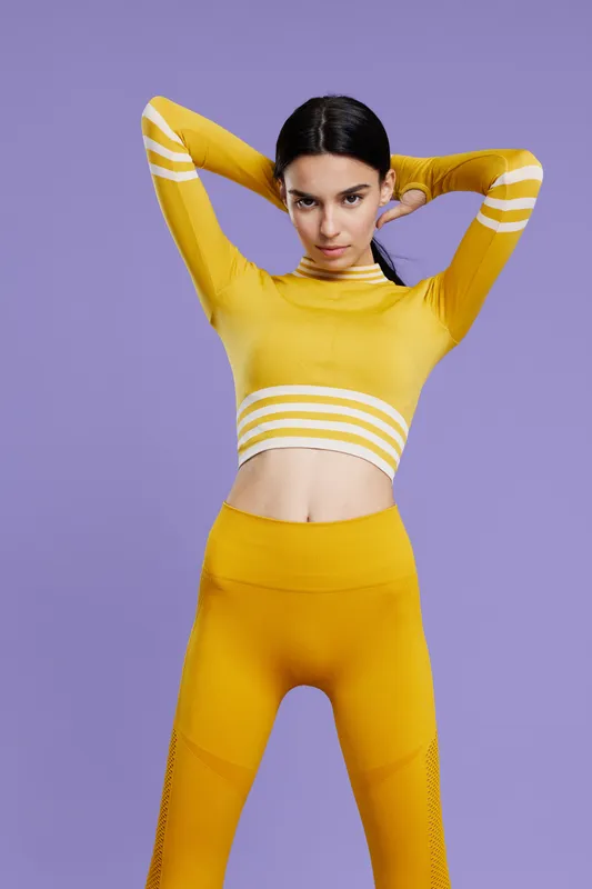 Woman sport fitness wearing yellow activewear stretches arms behind head against purple background, showing focus, strength, and healthy lifestyle.
