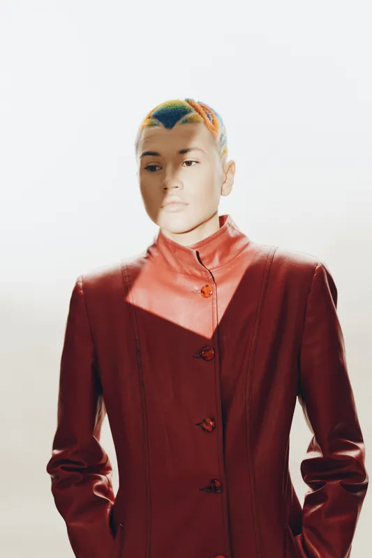 Woman with colorful short hair wearing a stylish red coat, standing in soft natural light against a white background, showcasing modern fashion and bold expression.