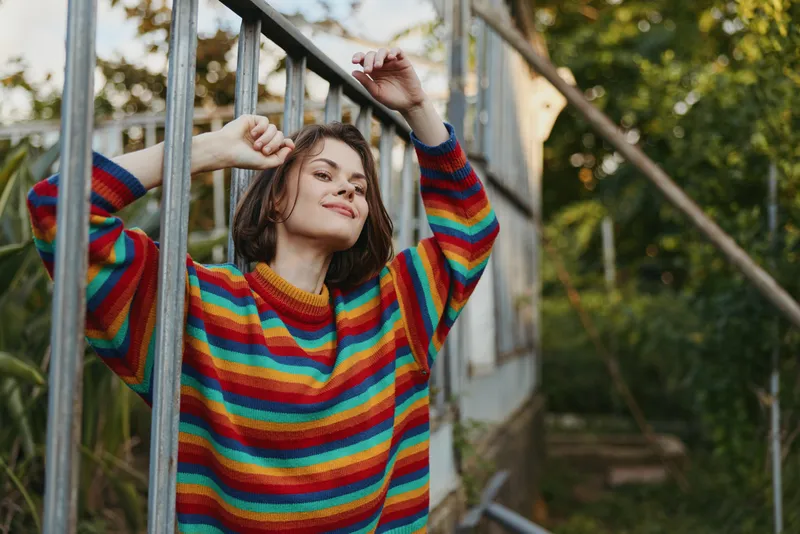Young woman in her twenties wearing a colorful striped sweater, smiling and leaning on a greenhouse frame in a lush outdoor garden. Casual outfit, short hair, relaxed lifestyle fashion portrait.
