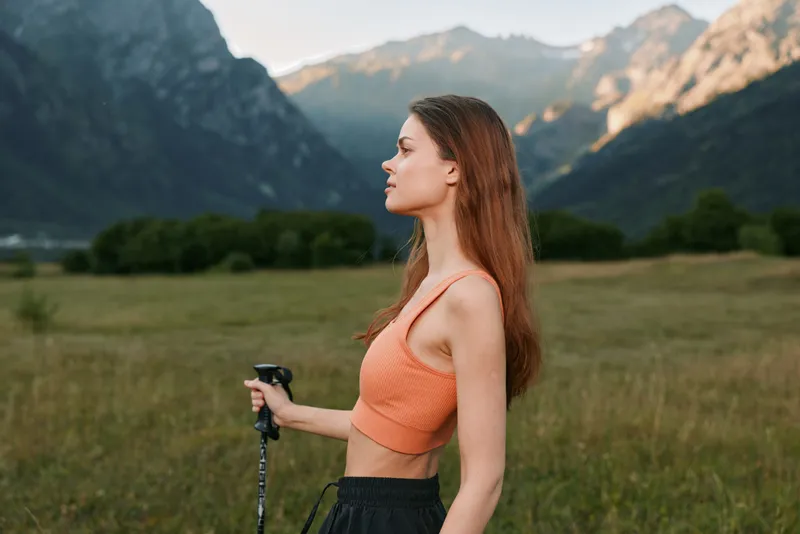 A fit female hiker stands in a scenic valley with distant mountains. She wears an orange sports top and holds a trekking pole, enjoying outdoor adventure and nature.