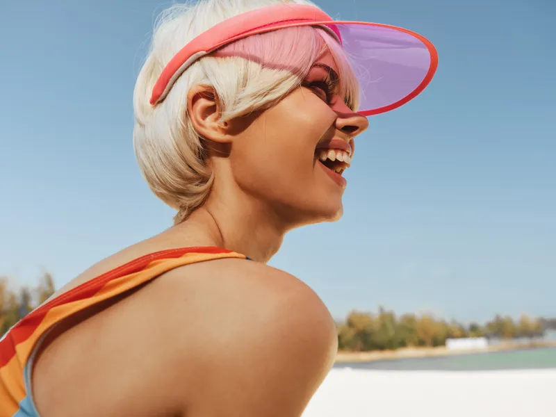 Close portrait of a joyful woman with short blonde hair wearing a pink visor, captured outdoors on a sunny day, conveying vibrant summer energy and chic playful style.