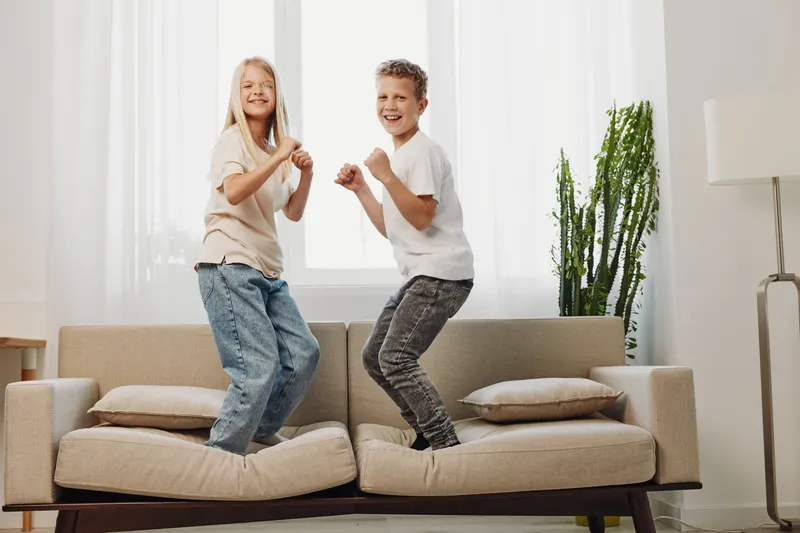 Happy children jumping on couch in living room, laughing and having fun together. Bright interior with natural light and modern furniture. Concept of playfulness, childhood joy and family time.