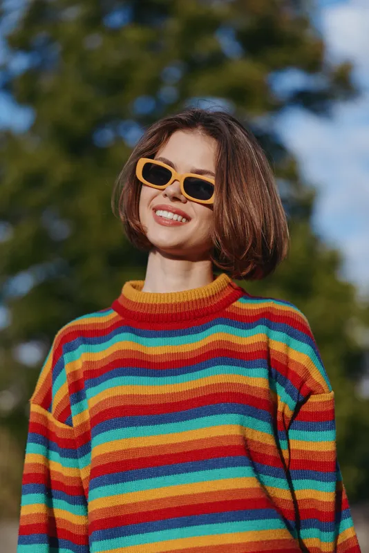 Woman smiling in a striped sweater and yellow sunglasses, short hair young adult outdoors in a portrait shot; colorful fashion casual outfit, joyful expression and sunny lifestyle mood.