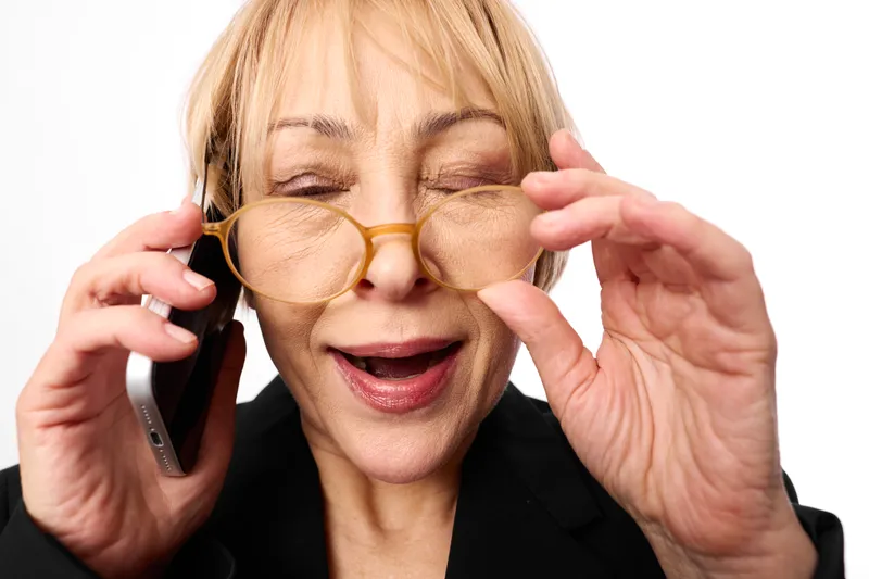 Elderly woman wearing gold glasses, smiling while holding frames with both hands, talking on smartphone, playful studio portrait, soft light, focused closeup, cheerful mood