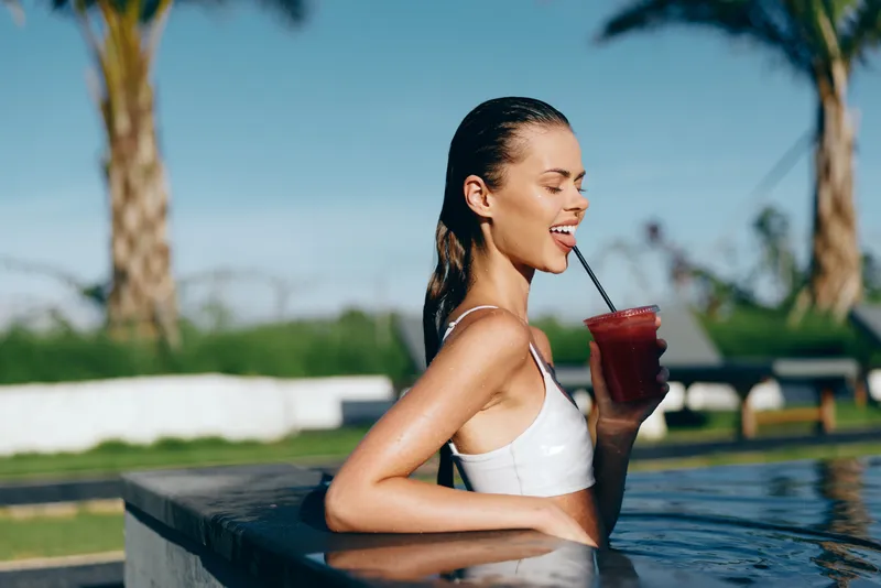 woman smiling pool poolside summer beverage drink hydration leisure lifestyle. Smiling woman in a white swimsuit relaxes by a bright pool, sipping a red beverage, with palm trees and sunlit water
