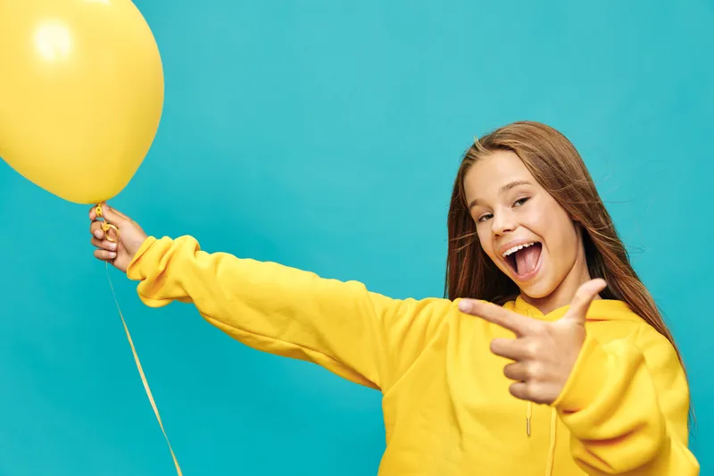 Girl in yellow sweatshirt holding yellow balloon and pointing finger at camera with happy expression on blue background smiling with excitement and joy.