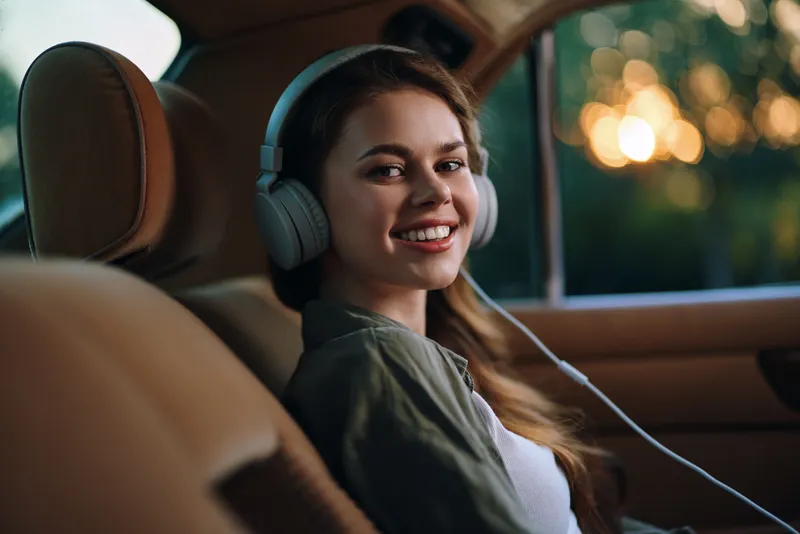 Young woman enjoying music in a car, smiling, wearing headphones, with a warm sunset background showcasing a relaxed and joyful mood.