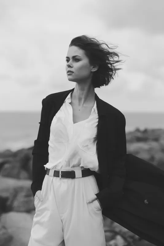 Fashionable woman outdoors wind swept wearing a blazer white shirt and light trousers monochrome scene with rugged coastline confident stance and modern street style