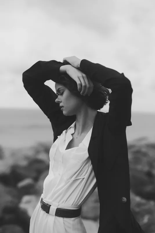 A stylish woman in a blazer and white shirt stands by the seaside, arms raised against the wind, producing a moody black and white fashion moment that conveys confidence, poise, and minimalist