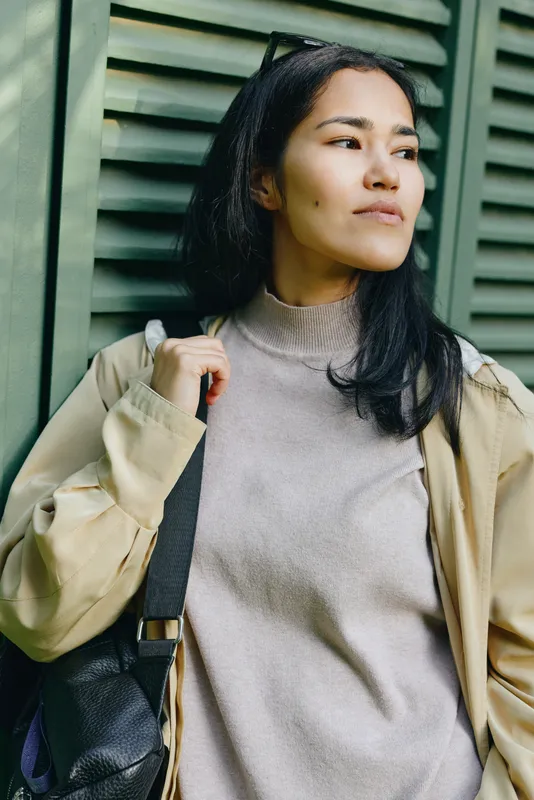 Woman, thoughtful and street style, stands near green shutters wearing a casual coat and soft knit sweater, carrying a bag, reflecting on daily life and urban fashion, captured in natural light with