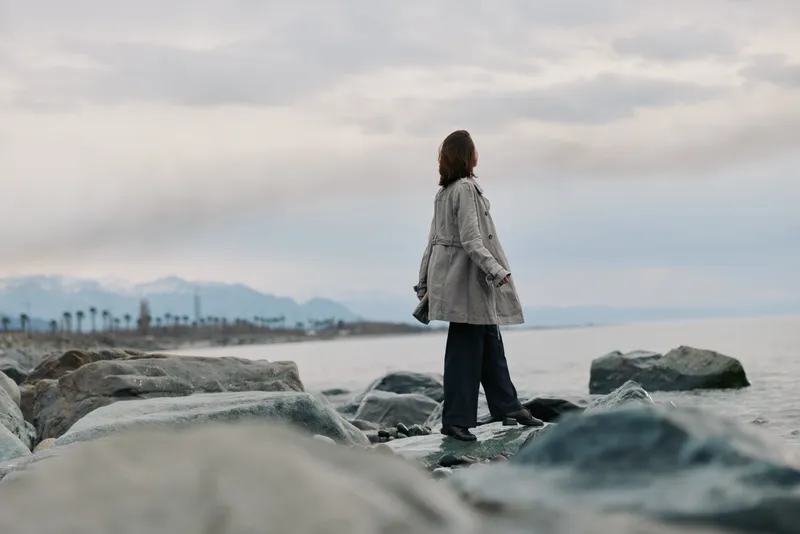 A woman in a checkered coat stands on rocks by a cloudy sea shore, looking into the distance. The scene evokes solitude, nature, and contemplation with muted colors and calm atmosphere.