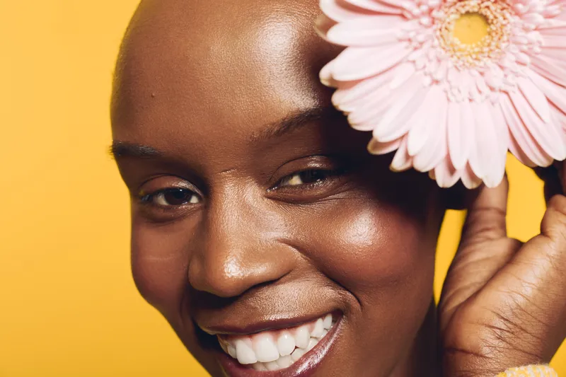 Close-up of a smiling black woman with dark skin holding a pink flower near her face on a yellow background. Concept of natural beauty, skincare, happiness, and wellness.