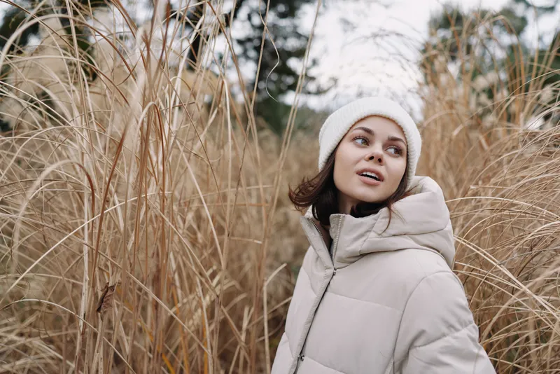 Woman in a light winter coat stands among tall dried grasses, gazing upward in a chilly outdoor setting, conveying calm, style and seasonal atmosphere in a natural field during soft daylight
