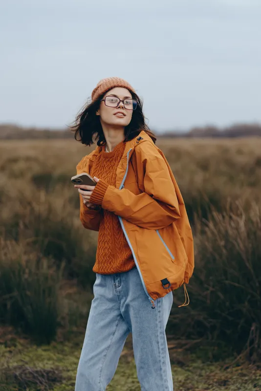 Woman lifestyle outdoors field portrait, wearing orange jacket, knit sweater, beanie and glasses, autumn daylight, walking with relaxed smile, denim jeans, soft wind, natural grassland background