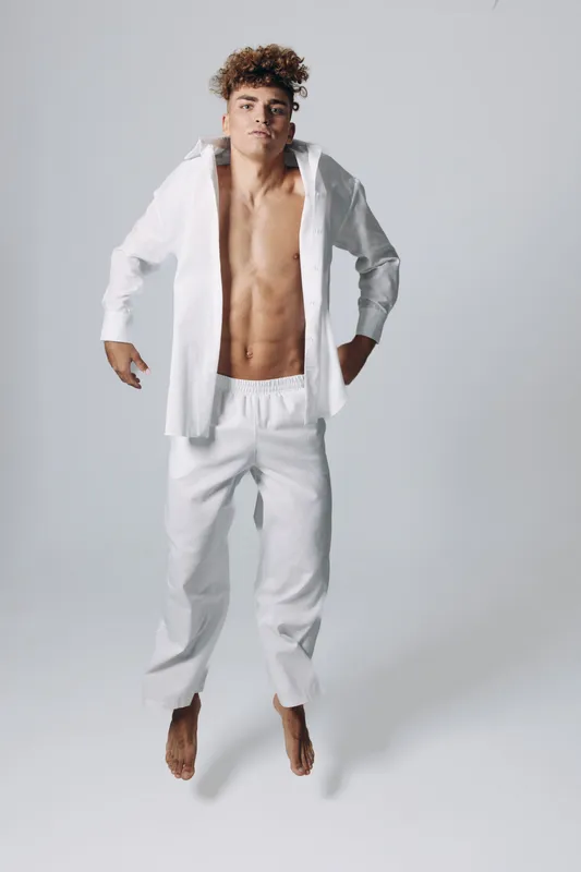 Young man jumping in white pants and open shirt with bare chest on plain background. Casual style, motion, fitness and confident expression in studio photo.