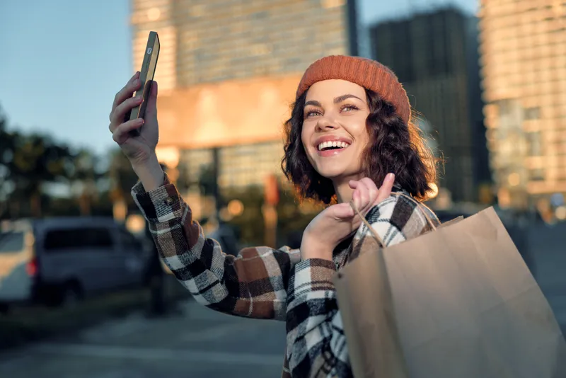 Woman shopping selfie smile urban bag beanie evening capturing candid lifestyle and authenticity in golden hour glow, emotional storytelling of mindful living during joyful city shopping.