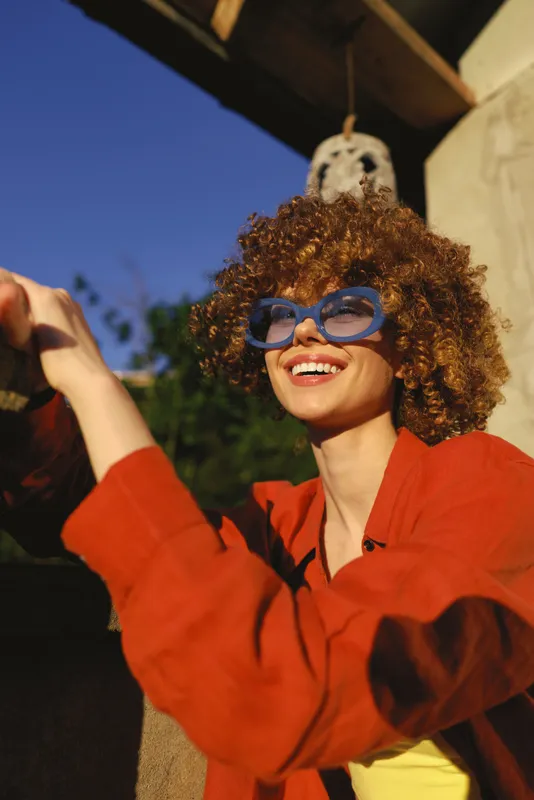Smiling woman with curly hair wearing a red jacket and blue glasses outdoors. Casual lifestyle portrait on a sunny day, capturing playful energy, confidence, and carefree style