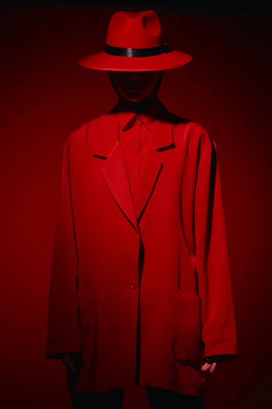 Mysterious model in oversized bright red blazer and wide-brimmed hat with black ribbon in dramatic red lighting. Concept for fashion, beauty, creative editorial, style.