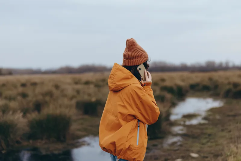 Woman lifestyle outdoor raincoat hat nature countryside river scene, young adult in mustard coat and knit hat, standing on a wet path, windy cloudy weather, carrying small dog