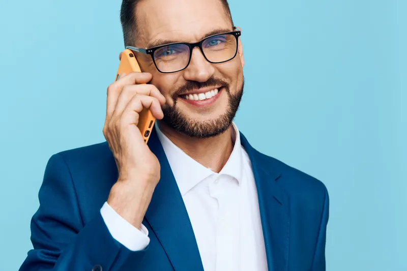 Professional male in a blue suit speaks on a mobile phone against an isolated blue background, showing confidence, warmth, and focus in a dynamic business moment.