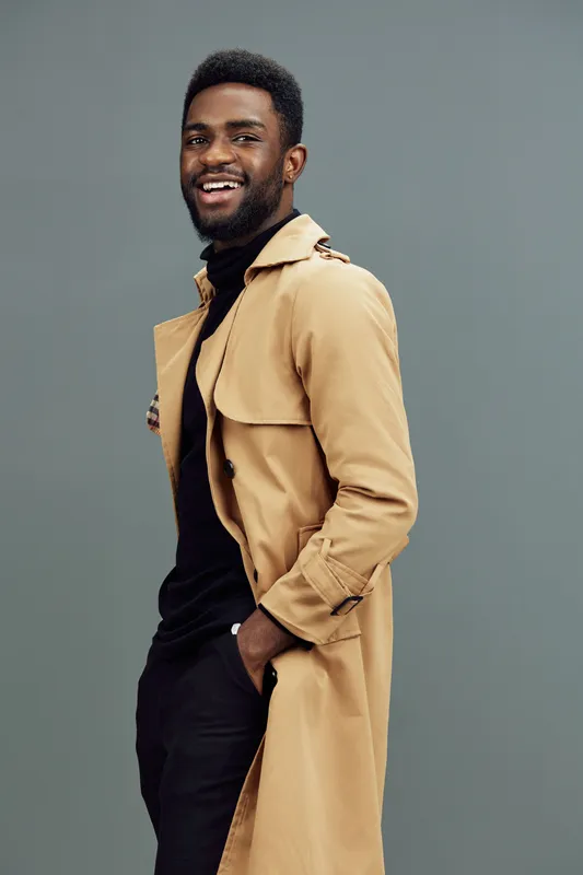 Smiling man in a stylish beige trench coat, exuding confidence and modern fashion against a muted grey background, perfect for lifestyle and fashion shoots
