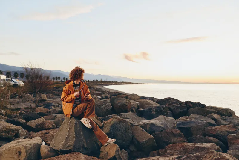 Woman lifestyle sunset sea rocks coast travel outdoor calm portrait, sitting on stone rocks near shoreline, orange coat and scarf, warm evening sunlight, peaceful relax