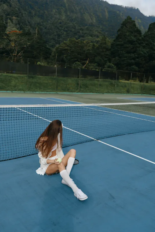 Young woman in sporty outfit sitting on tennis court, displaying a thoughtful emotion amidst a serene outdoor background