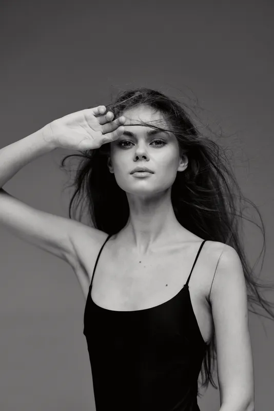 Black and white portrait of a young woman with long hair posing with hand near forehead in studio lighting