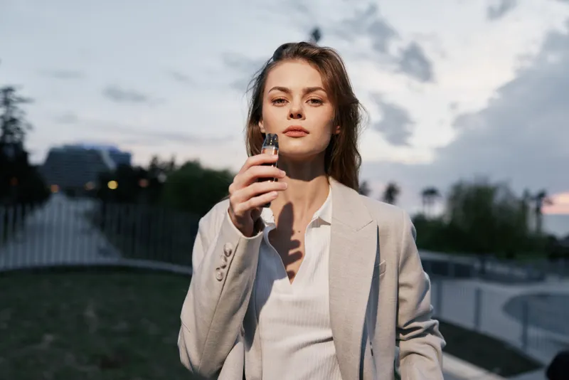 Confident woman in a blazer holding a smartphone outdoors, capturing a moment of empowerment at dusk with a modern backdrop.