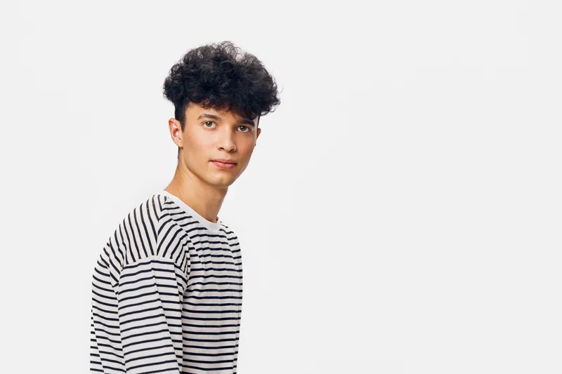 young man with curly hair wearing a striped shirt standing against white background expressing calm and casual style with natural lighting