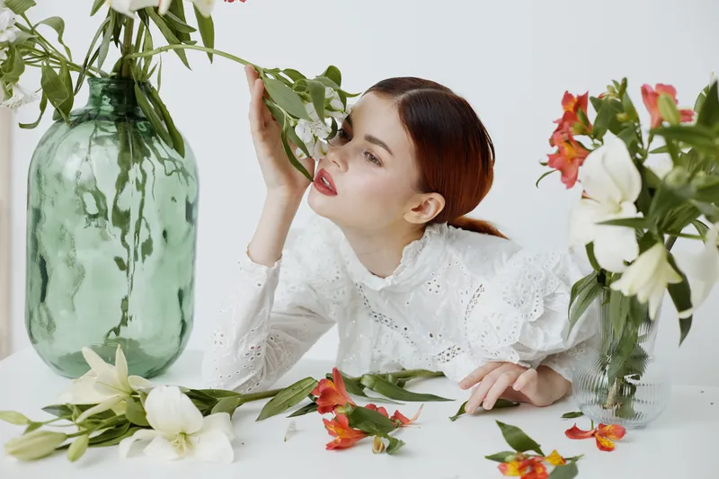 Young woman with red hair in a white lace blouse surrounded by fresh flowers, expressing tranquility and beauty in a soft pastel setting