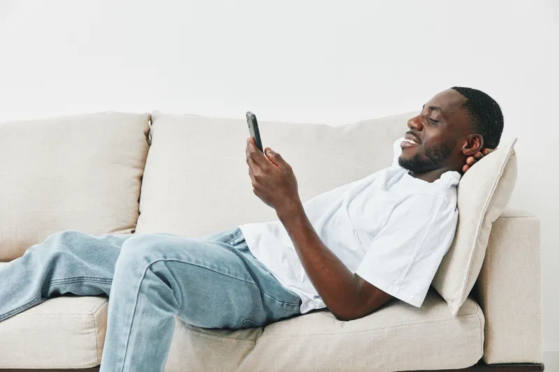 Relaxed young Black man using smartphone on a couch, showcasing modern lifestyle and comfort at home, with a casual outfit and joyful expression