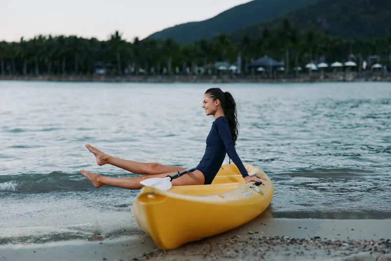 A woman sits in a bright yellow kayak on a calm shoreline, legs extended over the bow, gazing toward distant hills as gentle waves touch the sand under a muted sky.