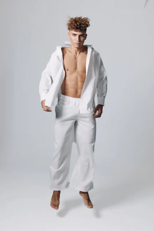 Young man with curly hair standing barefoot wearing loose white pants and open white shirt showing toned abs. Minimalist style and neutral background for fashion or health concept.