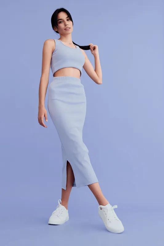 Young woman in a stylish blue crop top and matching skirt poses confidently against a pastel background, wearing casual white sneakers in a modern fashion look.