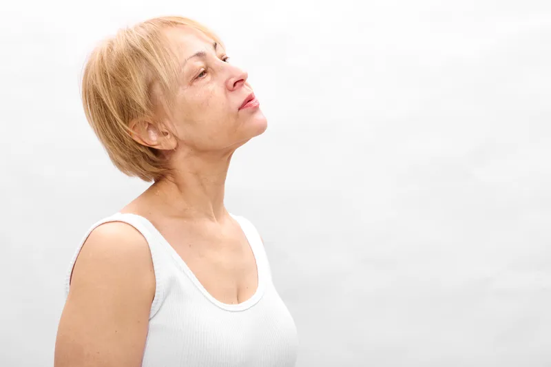 Elderly woman portrait in white tank top, head tilted upward, thoughtful face shows health and aging stress in bright studio light, neutral background, quiet worry and health check