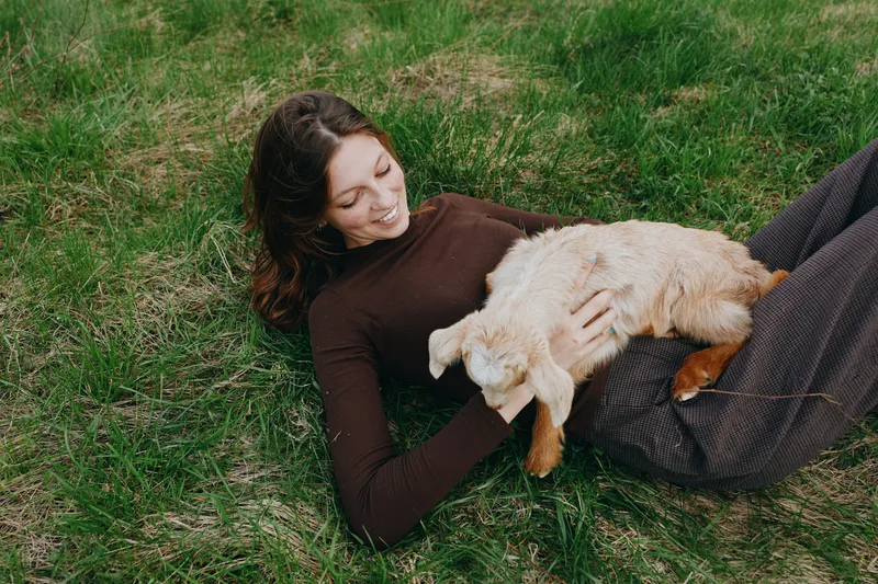 lifestyle, woman, goat, kid, outdoors, grass, cuddling, relaxation, a calm outdoor scene showing a woman lying on grass cradling a friendly goat kid in a tender moment