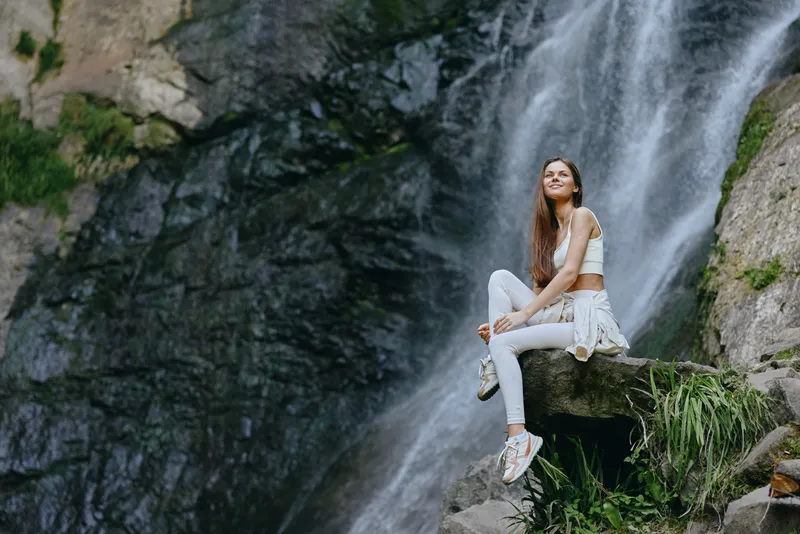 young woman sitting on rock near waterfall in nature, wearing white activewear, smiling with long hair, outdoor adventure and relaxation mood