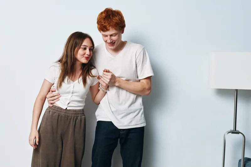 Couple standing close together against light wall, woman and man in casual clothes smiling and holding hands. Relationship, affection, friendship, cozy moment and relaxed mood indoors.