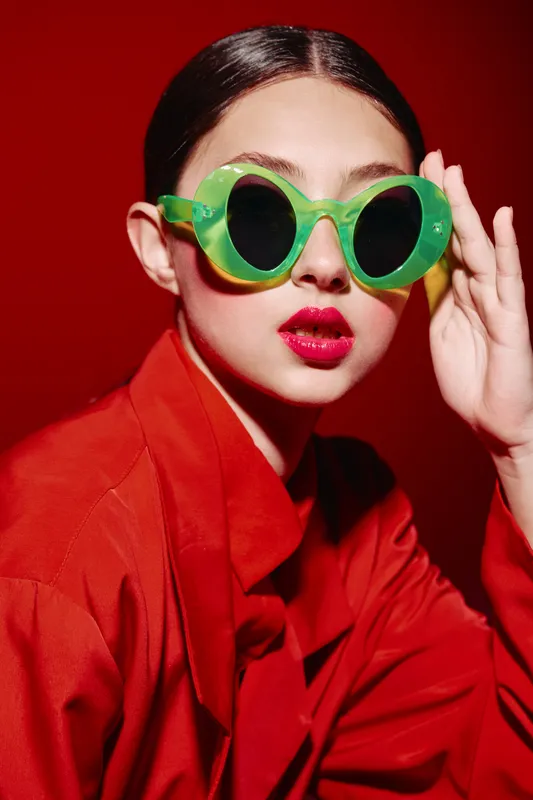 Stylish woman with bold red lips wearing vibrant green round sunglasses reacts confidently in bright red clothing on a red background. Concept of beauty fashion creativity.